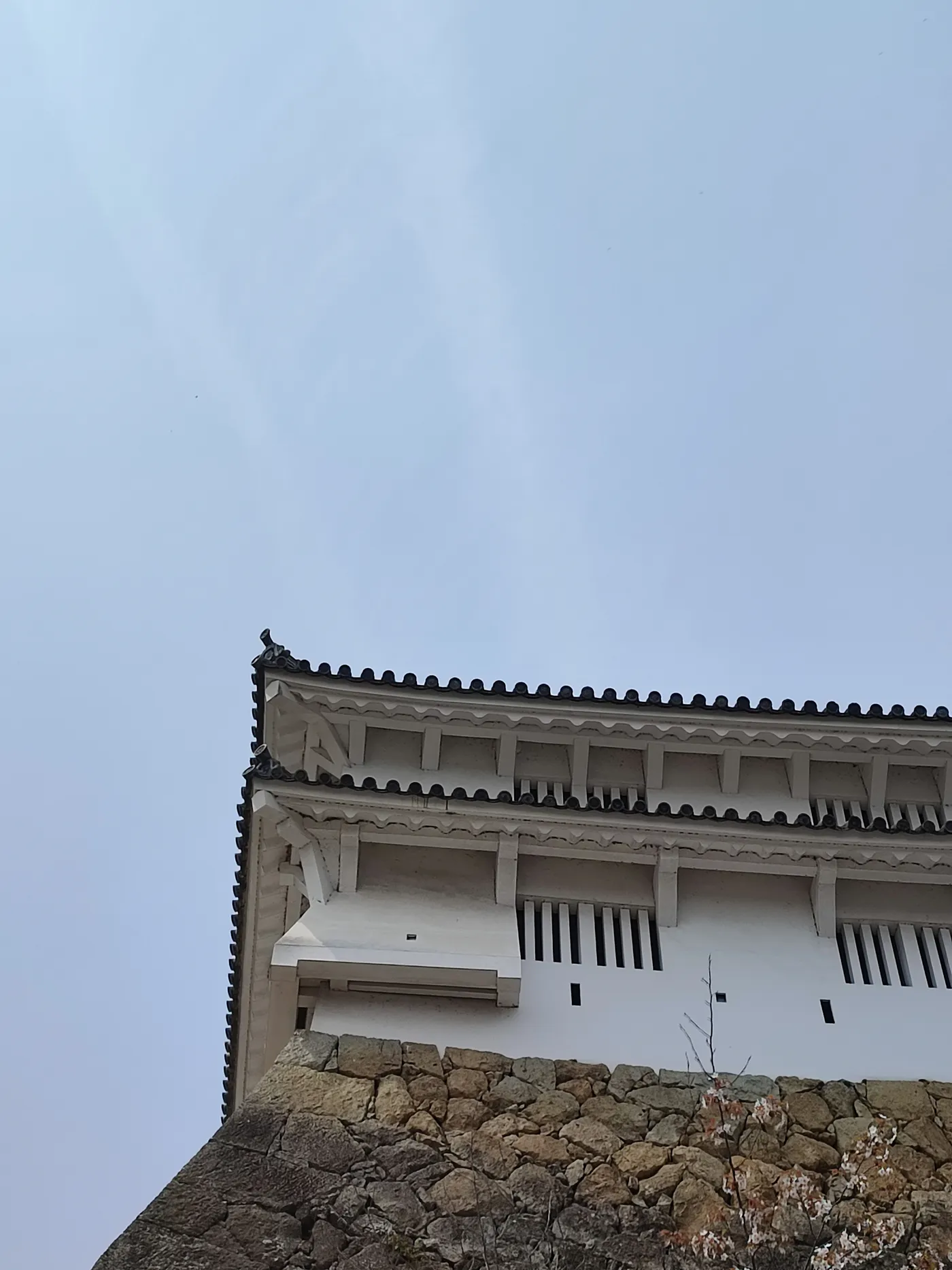 Corner of a white castle turret with tiled eaves and slatted windows, rising from a rough stone base against an overcast sky